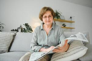 Senior senhora lendo dela jornal às casa relaxante em uma sofá e encarando sobre a topo às a espectador foto