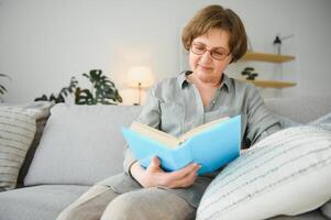 repousante leitura. retrato do pensativo envelhecido mulher lendo favorito literatura às acolhedor lar. ela é deitado em almofadas em confortável sofá. pernas estão coberto com suave cobertor. foto