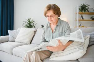 Senior senhora lendo dela jornal às casa relaxante em uma sofá e encarando sobre a topo às a espectador foto