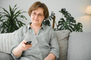 quebra notícias. chocado Senior mulher assistindo televisão às lar, sentado em sofá com controlo remoto controlador dentro mãos foto