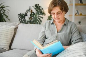 retrato do atraente focado alegre mulher sentado em divã lendo livro passatempo sonhar Acordado às casa casa plano interior. foto