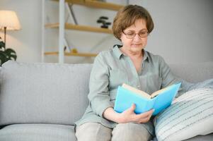 repousante leitura. retrato do pensativo envelhecido mulher lendo favorito literatura às acolhedor lar. ela é deitado em almofadas em confortável sofá. pernas estão coberto com suave cobertor. foto