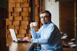 negócios, tecnologia e pessoas conceito , Senior homem de negocios com computador portátil computador bebendo café às moderno cafeteria. foto