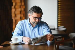 ativo Senior homem lendo jornal e bebendo café dentro restaurante foto