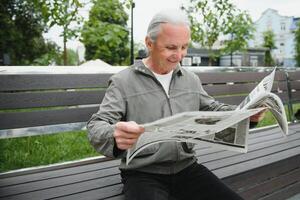 bonito avô senta em uma Banco dentro a parque e lê uma jornal. Senior grisalho cara. foto