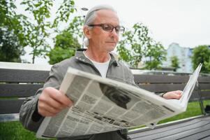 bonito avô senta em uma Banco dentro a parque e lê uma jornal. Senior grisalho cara. foto