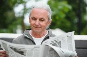 bonito avô senta em uma Banco dentro a parque e lê uma jornal. Senior grisalho cara. foto