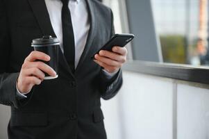 barbudo homem de negocios dentro formal terno em pausa usando Móvel telefone usar Smartphone. o negócio homem em pé lado de fora em moderno urbano cidade rua fundo com café copo dentro centro da cidade ao ar livre. cópia de espaço foto