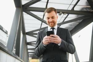 feliz sorridente homem de negocios vestindo Preto terno e usando moderno Smartphone perto escritório às cedo manhã, bem sucedido Empregador para faço uma acordo enquanto em pé perto arranha-céu escritório às noite, flare luz foto