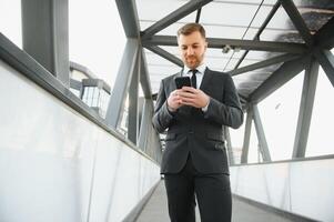 feliz sorridente homem de negocios vestindo Preto terno e usando moderno Smartphone perto escritório às cedo manhã, bem sucedido Empregador para faço uma acordo enquanto em pé perto arranha-céu escritório às noite, flare luz foto