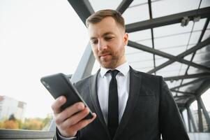 feliz sorridente homem de negocios vestindo Preto terno e usando moderno Smartphone perto escritório às cedo manhã, bem sucedido Empregador para faço uma acordo enquanto em pé perto arranha-céu escritório às noite, flare luz foto