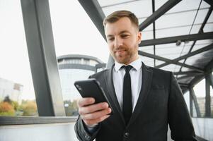 feliz sorridente homem de negocios vestindo Preto terno e usando moderno Smartphone perto escritório às cedo manhã, bem sucedido Empregador para faço uma acordo enquanto em pé perto arranha-céu escritório às noite, flare luz foto