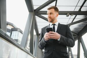 feliz sorridente homem de negocios vestindo Preto terno e usando moderno Smartphone perto escritório às cedo manhã, bem sucedido Empregador para faço uma acordo enquanto em pé perto arranha-céu escritório às noite, flare luz foto