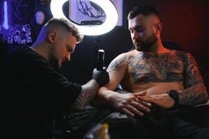 barbudo tatuagem artista trabalhando às dele estúdio tatuagem manga em a braço do dele masculino cliente. homem obtendo tatuado de profissional tatuador foto