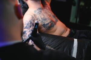 masculino tatuagem artista segurando uma tatuagem pistola, mostrando uma processo do fazer tatuagens em uma masculino tatuado do modelo braço foto