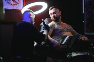 barbudo tatuagem artista trabalhando às dele estúdio tatuagem manga em a braço do dele masculino cliente. homem obtendo tatuado de profissional tatuador foto
