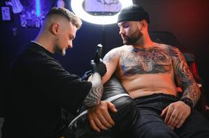 barbudo tatuagem artista trabalhando às dele estúdio tatuagem manga em a braço do dele masculino cliente. homem obtendo tatuado de profissional tatuador foto