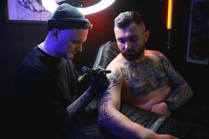 masculino tatuagem artista segurando uma tatuagem pistola, mostrando uma processo do fazer tatuagens em uma masculino tatuado do modelo braço foto