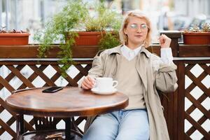 jovem à moda lindo mulher sentado dentro cidade cafeteria dentro rua, Primavera moda tendência estilo, bebendo café com leite, Rosa bolsa, saiu, emocional, feliz, sorridente foto