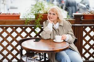 jovem à moda lindo mulher sentado dentro cidade cafeteria dentro rua, Primavera moda tendência estilo, bebendo café com leite, Rosa bolsa, saiu, emocional, feliz, sorridente foto