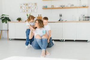 feliz jovem família, mãe com pequeno crianças sentar em caloroso de madeira chão dentro Novo moderno Projeto cozinha, mãe com animado pequeno crianças relaxar descansar dentro próprio renovado apartamento, comovente conceito foto