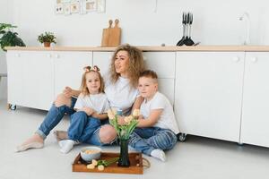 feliz mãe abraçando filha e filho sentado em de madeira chão dentro moderno cozinha às lar. mãe abraçando crianças. feliz família relação conceito foto