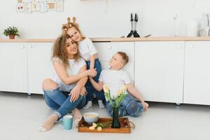 feliz jovem família, mãe com pequeno crianças sentar em caloroso de madeira chão dentro Novo moderno Projeto cozinha, mãe com animado pequeno crianças relaxar descansar dentro próprio renovado apartamento, comovente conceito foto