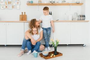 feliz mãe abraçando filha e filho sentado em de madeira chão dentro moderno cozinha às lar. mãe abraçando crianças. feliz família relação conceito foto