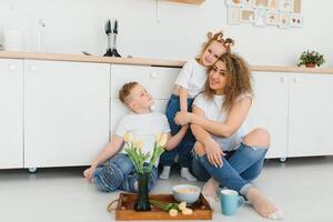 feliz jovem família, mãe com pequeno crianças sentar em caloroso de madeira chão dentro Novo moderno Projeto cozinha, mãe com animado pequeno crianças relaxar descansar dentro próprio renovado apartamento, comovente conceito foto