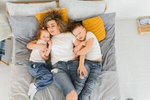 retrato do sorridente jovem mamãe abraçando crianças pequeno filho e filha. feliz família. mãe com crianças sentado em cama e amoroso a Boa vezes junto. fechar-se foto