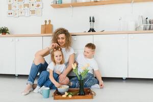 feliz mãe abraçando filha e filho sentado em de madeira chão dentro moderno cozinha às lar. mãe abraçando crianças. feliz família relação conceito foto