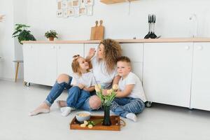 feliz jovem família, mãe com pequeno crianças sentar em caloroso de madeira chão dentro Novo moderno Projeto cozinha, mãe com animado pequeno crianças relaxar descansar dentro próprio renovado apartamento, comovente conceito foto