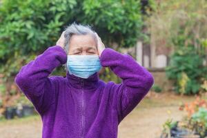 uma Senior mulher vestindo uma face mascarar tem uma dor de cabeça enquanto em pé dentro uma jardim. mascarar para proteção COVID-19, vírus, bactérias, etc. conceito do envelhecido pessoas e cuidados de saúde foto