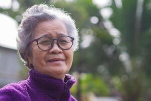 retrato do uma Senior mulher com curto cinzento cabelo vestindo copos, sorridente, e olhando às a Câmera enquanto em pé ao ar livre. espaço para texto. conceito do envelhecido pessoas e cuidados de saúde foto