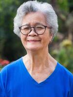 uma retrato do uma Senior mulher com curto cinzento cabelo, vestindo copos, sorridente, e olhando às a Câmera enquanto em pé dentro uma jardim. espaço para texto. conceito do envelhecido pessoas e cuidados de saúde foto