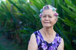 retrato do uma Senior mulher com cabelo rolos, braços cruzado, sorridente, e olhando às a Câmera enquanto em pé dentro uma jardim. espaço para texto. conceitos do envelhecido pessoas e cuidados de saúde foto