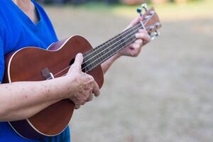 fechar-se do mãos idosos mulher jogando ukulele dentro dela jardim. relaxante de jogar pequeno guitarra feliz. seletivo foco. conceito do velho pessoas e saúde Cuidado foto