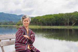 alegre idosos ásia mulher braços cruzado, sorridente, e olhando às a Câmera enquanto sentado em uma de madeira cadeira ao lado a lago. espaço para texto. conceito do envelhecido pessoas e relaxamento foto