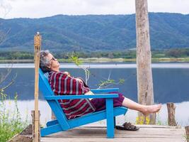 Senior mulher vestindo oculos de sol, braços cruzado enquanto sentado em uma de madeira cadeira lado do a lago. espaço para texto. conceito do envelhecido pessoas e relaxamento foto