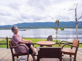 retrato do Senior ásia mulher vestindo óculos olhando longe enquanto sentado em uma de madeira cadeira lado a lago. conceito do envelhecido pessoas e relaxamento foto