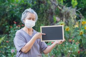 retrato do a idosos mulher segurando uma quadro-negro e vestindo uma face mascarar para protegendo vírus, COVID-19, coronavírus, bactérias, pólen grãos, ar poluição PM 2.5, e mais. cuidados de saúde conceito foto