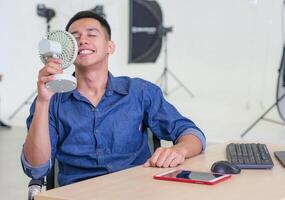 jovem homem segurando uma pequeno plástico ventilador sopro para dele face Porque do quente clima enquanto sentado em uma cadeira dentro a escritório. ele é olhos fechados e sorridente com relaxamento. estúdio local de trabalho foto