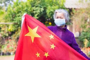Senior mulher vestindo uma cirúrgico mascarar e segurando a China bandeira para dando encorajamento para a chinês para luta covid-19 ou coronavírus. conceito do envelhecido pessoas e cuidados de saúde foto