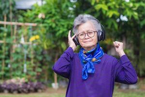 alegre Senior mulher com curto cinzento cabelo, vestindo fones de ouvido ouvindo para uma favorito canção, sorridente e olhando às a Câmera enquanto em pé dentro uma jardim. conceito do envelhecido pessoas e relaxamento foto