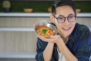 jovem do homem vestindo óculos sentado olhando às a Câmera, sorridente e mostrando a frito ovo com colorida coberturas servido em uma frigideira. simples Comida para café da manhã. foto