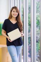 retrato do uma jovem mulher vestindo Preto Camisetas segurando uma parcela caixa, sorridente e olhando às a Câmera enquanto em pé dentro a escritório. conceito do o negócio e comércio eletrônico foto