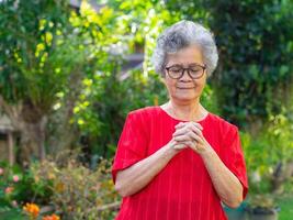 Senior mulher com curto branco cabelo vestindo copos, Rezar, e fechadas olhos enquanto em pé dentro a jardim. espaço para texto. conceito do envelhecido pessoas e cuidados de saúde foto