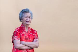 Senior mulher vestindo uma tradicional cheongsam qipao vestir, braços cruzado, sorridente, e olhando às a Câmera enquanto em pé em luz laranja fundo. chinês Novo ano conceito foto