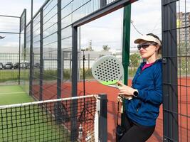 retrato do positivo jovem mulher com raquete e padel bola em tênis quadra foto