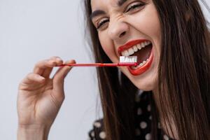 engraçado menina com vermelho lábios mantém escova de dente entre saudável branco dentes enquanto posando às a estúdio às a branco fundo. oral Cuidado conceito foto